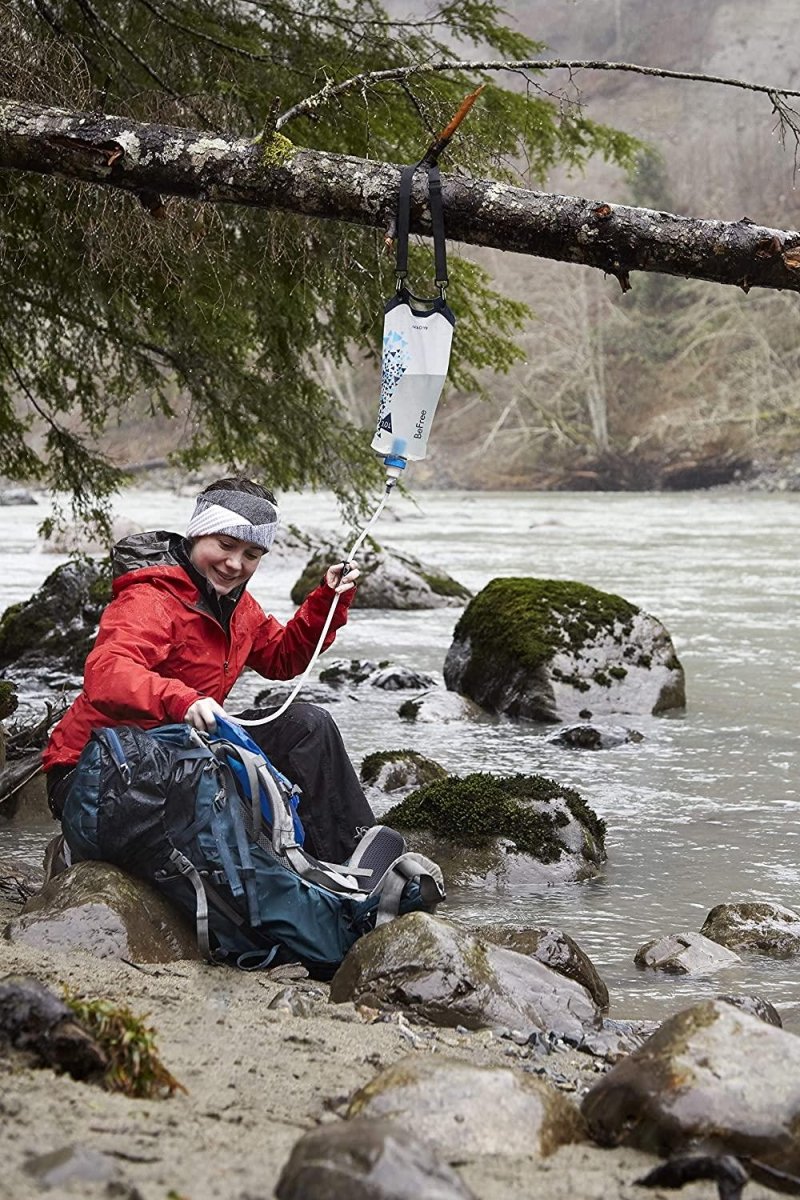 Person in red jacket using Katadyn BeFree Gravity 3L water filter hanging from tree branch by river outdoors