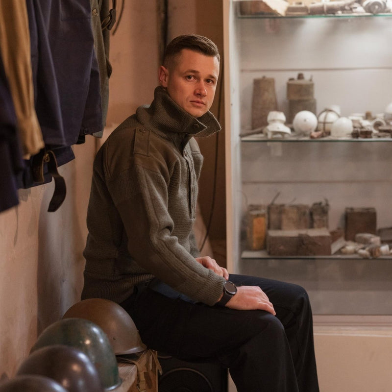 Man wearing an olive Austrian Army Alpine wool pullover sweater with high collar sitting indoors near military helmets and artifacts