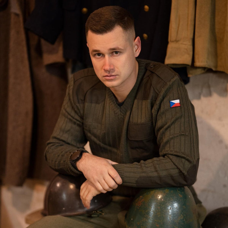 Man wearing original olive drab Czech army wool sweater with V-neck and flag patch on sleeve, sitting with helmets.